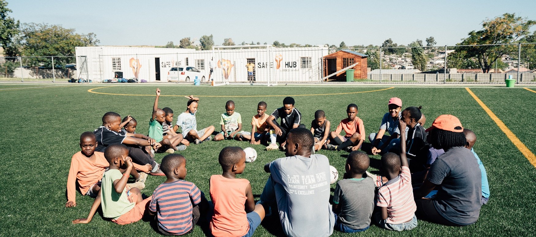 dunkelhäutige Kinder sitzen im Kreis auf einem Fußballfeld