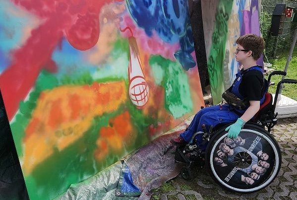 Wandmalerei auf Mauer, davor Person in Rollstuhl