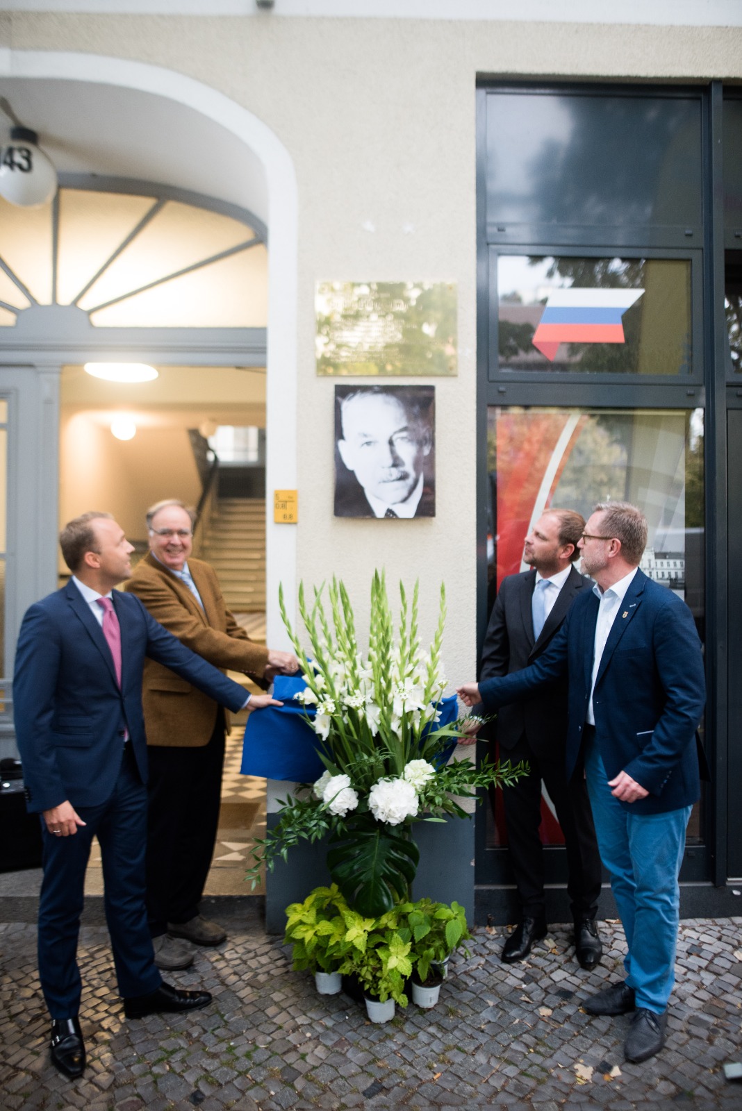 Vier Männer schauen zu Gedenktafel an Hauswand, davor großer Blumenstrauß